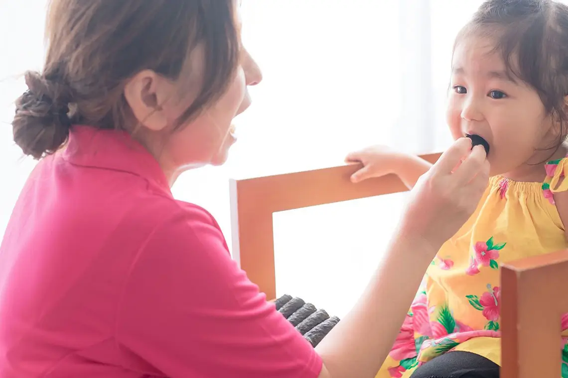 鮮やかなピンク色の服を着た母親が、花柄の黄色いワンピース姿の子供へ優しく食事を介助しています。
