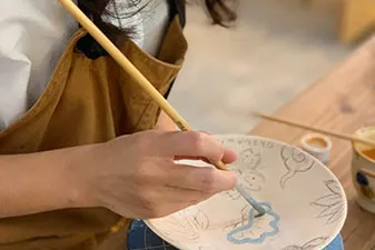 エプロン姿の人物が細い筆を使い、素焼きの白い皿に淡いブルーの絵具で模様を描いています。