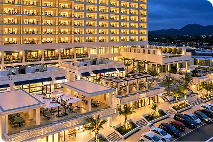 夜の帳が下りる中、温かな光を放つホテルの高層棟と、開放的なテラスが広がるオキナワ ハナサキマルシェの全景です。