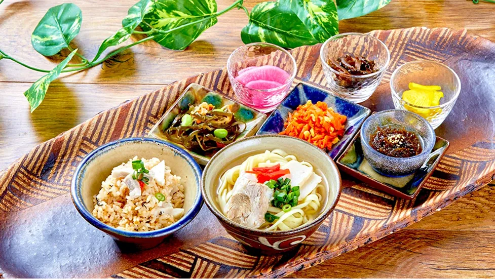 網目模様が刻まれた長い木のプレートに、沖縄そばや炊き込みご飯、色鮮やかな小鉢が美しく並べられています。