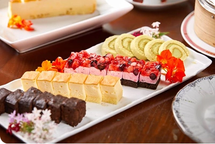 細長くカットされたチョコやチーズケーキ、ベリーのムースが白い長方形の皿に整然と並び、鮮やかなエディブルフラワーが彩りを添えています。