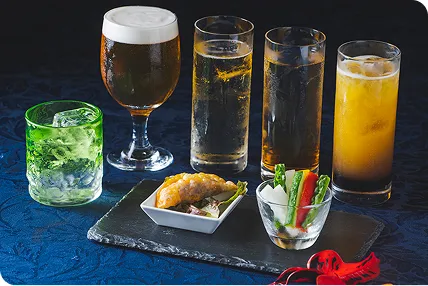 濃い青色の布が敷かれたテーブルに、泡立つビールや色鮮やかなカクテルと、黒いプレートに盛られた軽食が並んでいます。