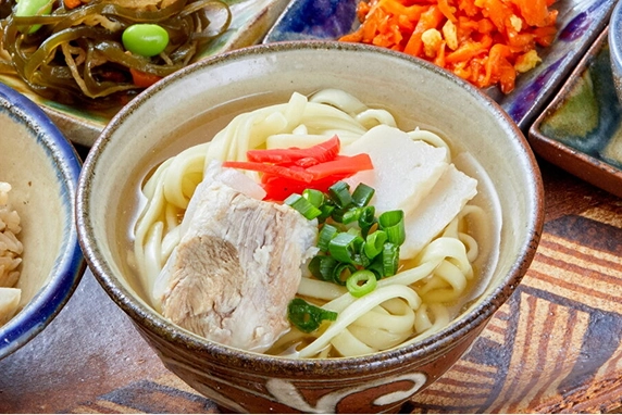 陶器の椀に入った沖縄そばは、透き通った出汁に平打ち麺が浸かり、肉厚な三枚肉と刻みネギ、紅生姜が彩りを添えています。