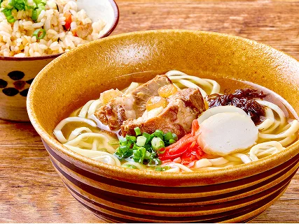 琥珀色の器に盛られた沖縄そばには三枚肉や紅生姜が乗り、傍らに炊き込みご飯の小鉢が添えられています。