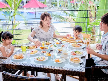 ヤシの木が茂る南国のテラスで、家族が色とりどりの料理が並ぶビュッフェの朝食を楽しんでいます。
