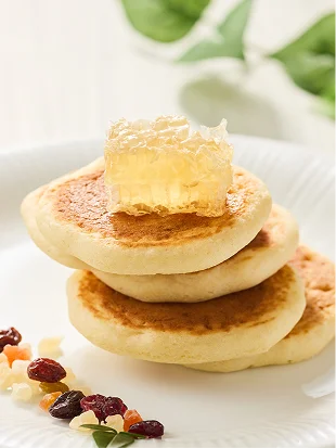 白い皿に重ねた焼きたてのパンケーキの上で、半透明の巣蜜が輝き、傍らに刻んだドライフルーツが添えられています。