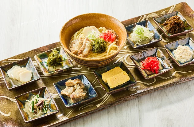 茶褐色の長皿に沖縄そばと小鉢が並び、三枚肉や紅生姜などの薬味が華やかに彩っています。