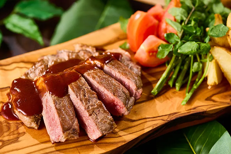オリーブの木目が浮かぶプレートに、焼き色がついたステーキと瑞々しいトマト、ハーブが添えられています。
