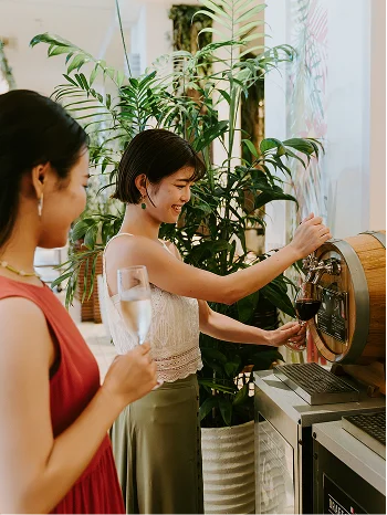 観葉植物が並ぶ店内で、笑顔の女性が木樽のサーバーからワインをグラスへ注いでいます。