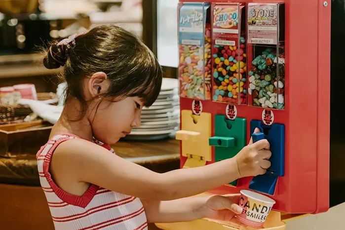 赤色の筐体のキャンディサーバーから、少女が青いレバーを引いてカラフルなお菓子をカップへ注いでいます。