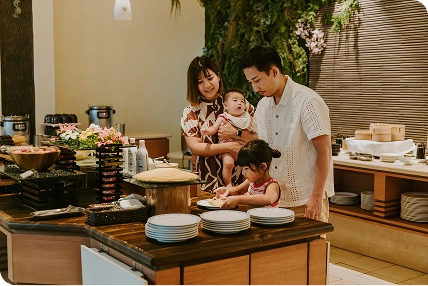 家族連れがビュッフェレストラン「マーセン」のカウンターで料理を選んでおり、背景にはやんばるの森を想起させる緑が配置されています。