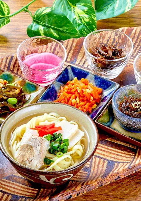 沖縄そばの丼を囲むように、赤や茶の小鉢に入った沖縄料理が幾何学模様の木製トレイへ配置されています。