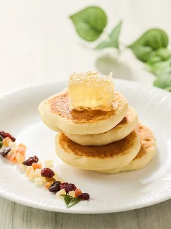 白い皿に積み重ねたパンケーキの頂点に琥珀色の巣蜜がのり、周囲に刻んだドライフルーツが彩りを添えています。