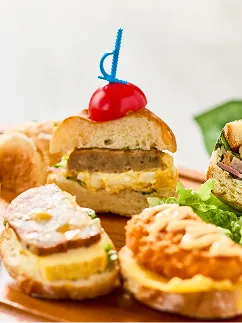 青いピックを刺したミニバーガーや揚げ物をのせたバゲットが、滑らかな木製ボードの上に並んでいます。