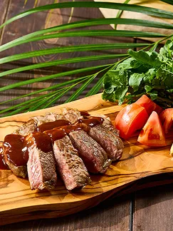 オリーブの木目が鮮やかなプレートに、ソースを纏った焼きステーキと瑞々しいトマトが盛り付けられています。