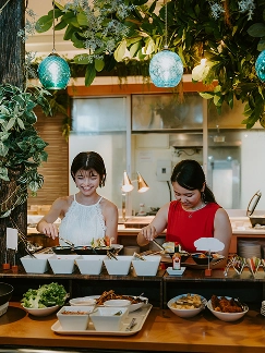 やんばるの森を模した装飾の下、二人の客がビュッフェカウンターでタコスや和洋食の料理をトングで選んでいます。