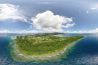 深い藍色の外洋に囲まれた緑豊かな島が、透明度の高い浅瀬と白い波打ち際を伴って本部半島を背景に浮かんでいます。