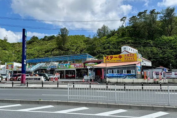 青空の下、黄色い看板が目を引く「道の駅許田」の施設が、緑に覆われた山並みを背景に並んでいます。